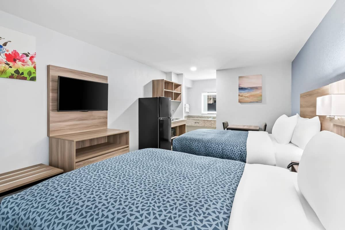 A modern hotel room with two beds, a flat-screen TV, a refrigerator, a wooden cabinet, and a small kitchenette.