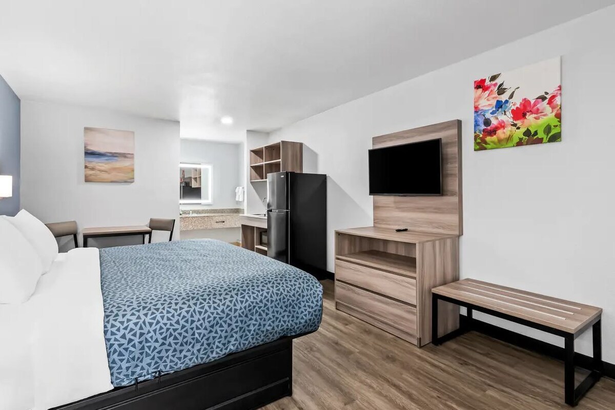A modern hotel room with a bed, a desk, a refrigerator, a bench, and a TV mounted on the wall.