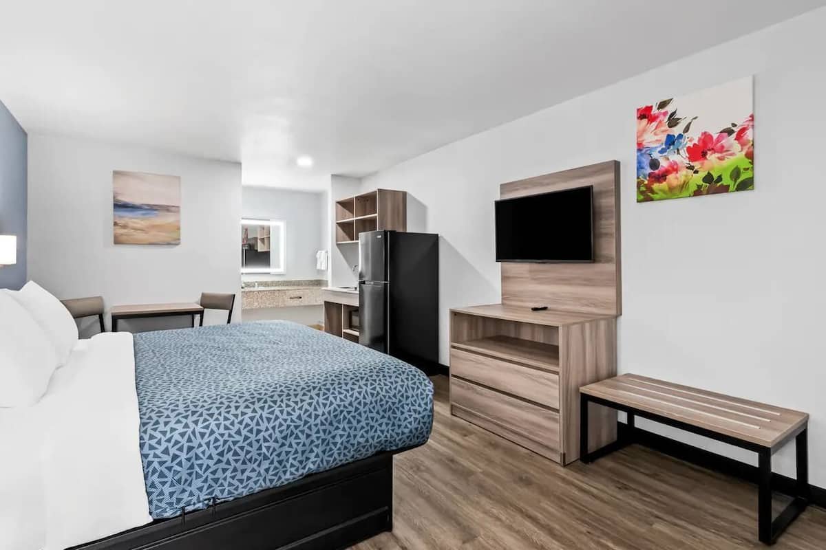 A modern hotel room with a bed, a desk, a refrigerator, a bench, and a TV mounted on the wall.