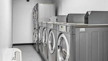 Een wasserette met meerdere wasmachines en drogers, een ventilatie-unit en een witte opbergwagen.