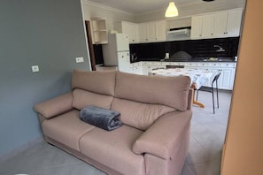 Een woonkamer met een beige bank, een eettafel met stoelen, en een keuken met witte kasten en een zwarte achterwand.