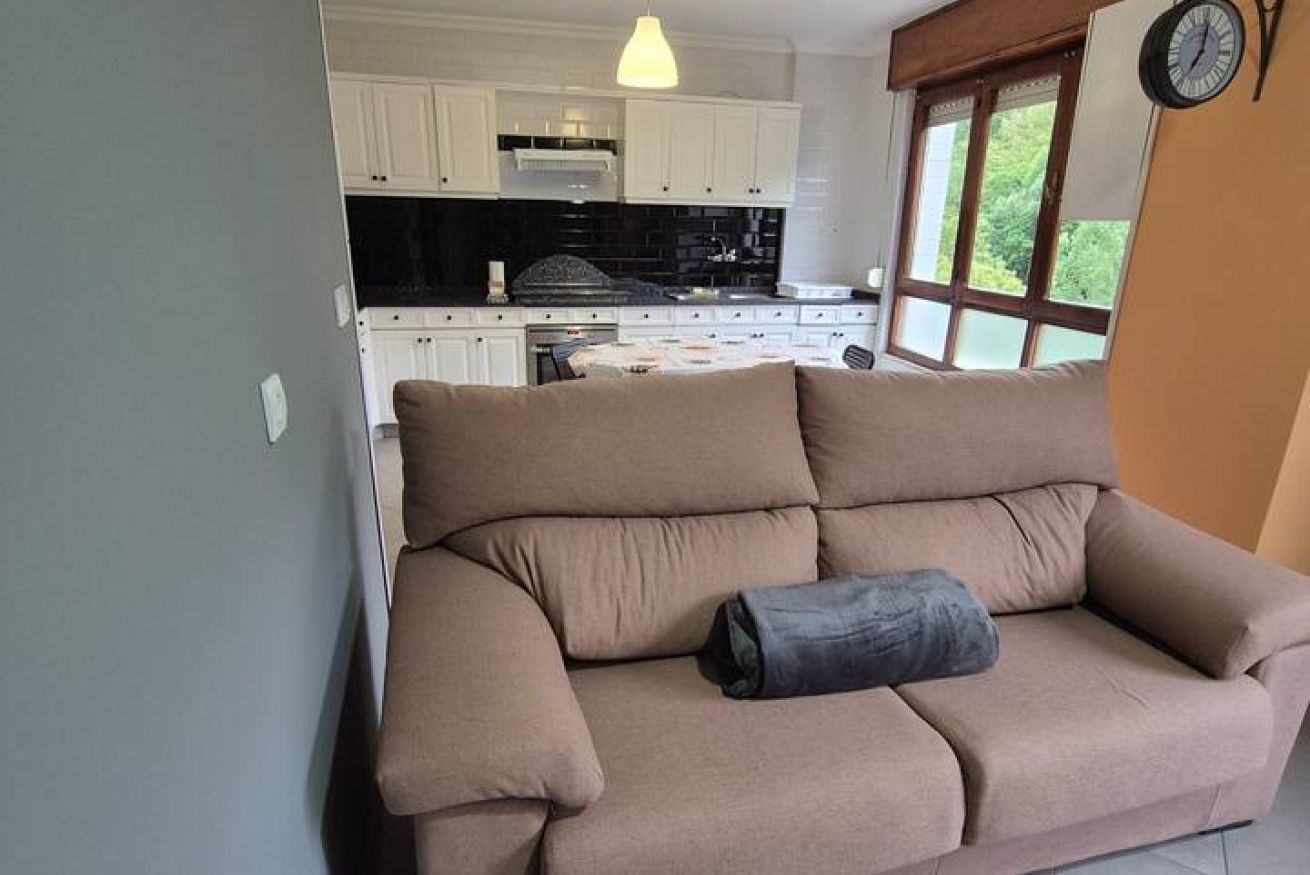 Una sala de estar con un sofá color crema, una ventana con vistas, y una cocina con armarios blancos y encimera negra.