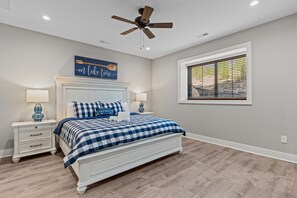 A bedroom with a bed, two bedside tables, a ceiling fan, a window with blinds, and a wall sign.