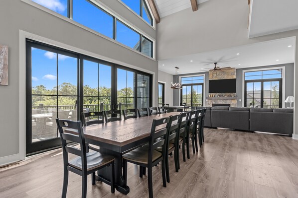 A spacious dining area with a long table and chairs, large windows, and a modern kitchen with a stone fireplace.
