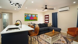 Ein modernes Wohnzimmer mit einem grauen Sofa, orangefarbenen Sesseln, einem Holztisch und einem farbenfrohen Gemälde an der Wand.