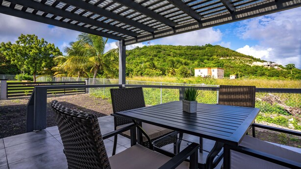 Eine Außenpatio mit Tisch und Stühlen, einer Pergola darüber und Blick auf einen grünen Hügel mit Gebäuden in der Ferne.