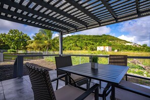 An outdoor patio with a table and chairs, a pergola overhead, and a view of a green hillside with buildings in the distance. - Modern Comfort Near Beach – Family-Friendly Stay (Osbourn)