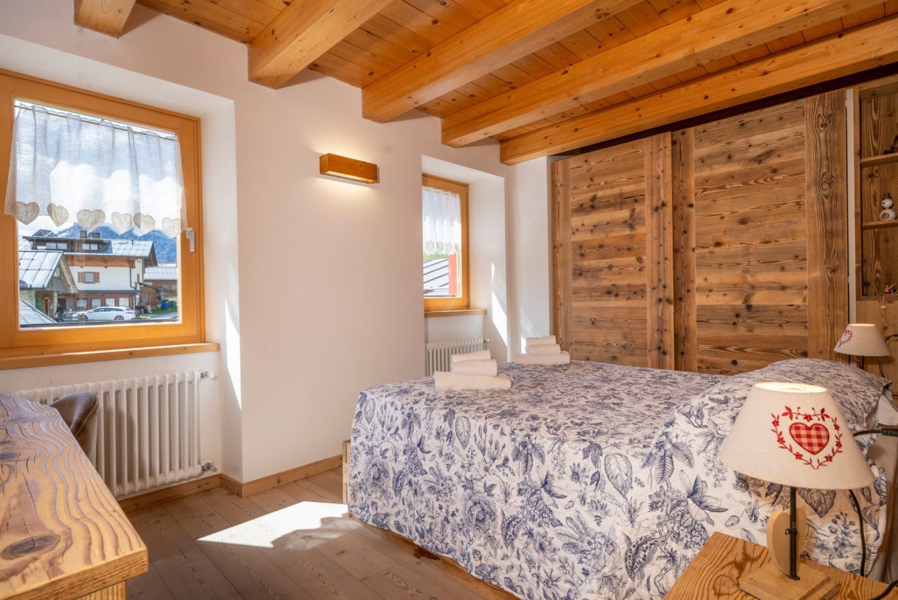 A bedroom with a wooden ceiling, a bed with a floral bedspread, a window with a view of a house, and a wooden wardrobe.
