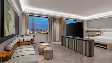 A modern hotel room with a large TV, a sofa, a dining table, and a view of the city at dusk.