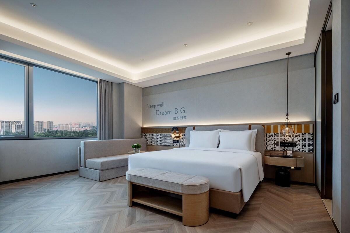 Habitación de hotel moderna con una cama grande, un banco, una mesita de noche y vistas a la ciudad.