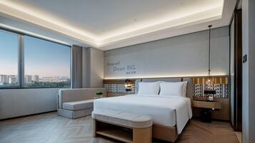 A modern hotel room with a large bed, a bench, a nightstand, and a view of the city.