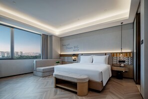 A modern hotel room with a large bed, a bench, a nightstand, and a view of the city.
