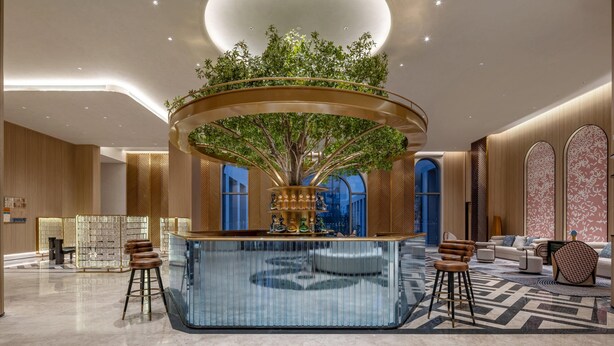 Eine moderne Lobby mit einer zentralen, runden Bar, einem großen Baum und einer Sitzgruppe mit Barhocker.