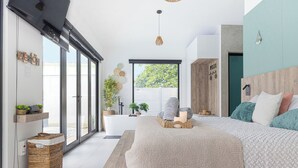 Une chambre moderne avec un grand lit, une tête de lit en bois et une vue sur l’extérieur grâce à de grandes fenêtres.