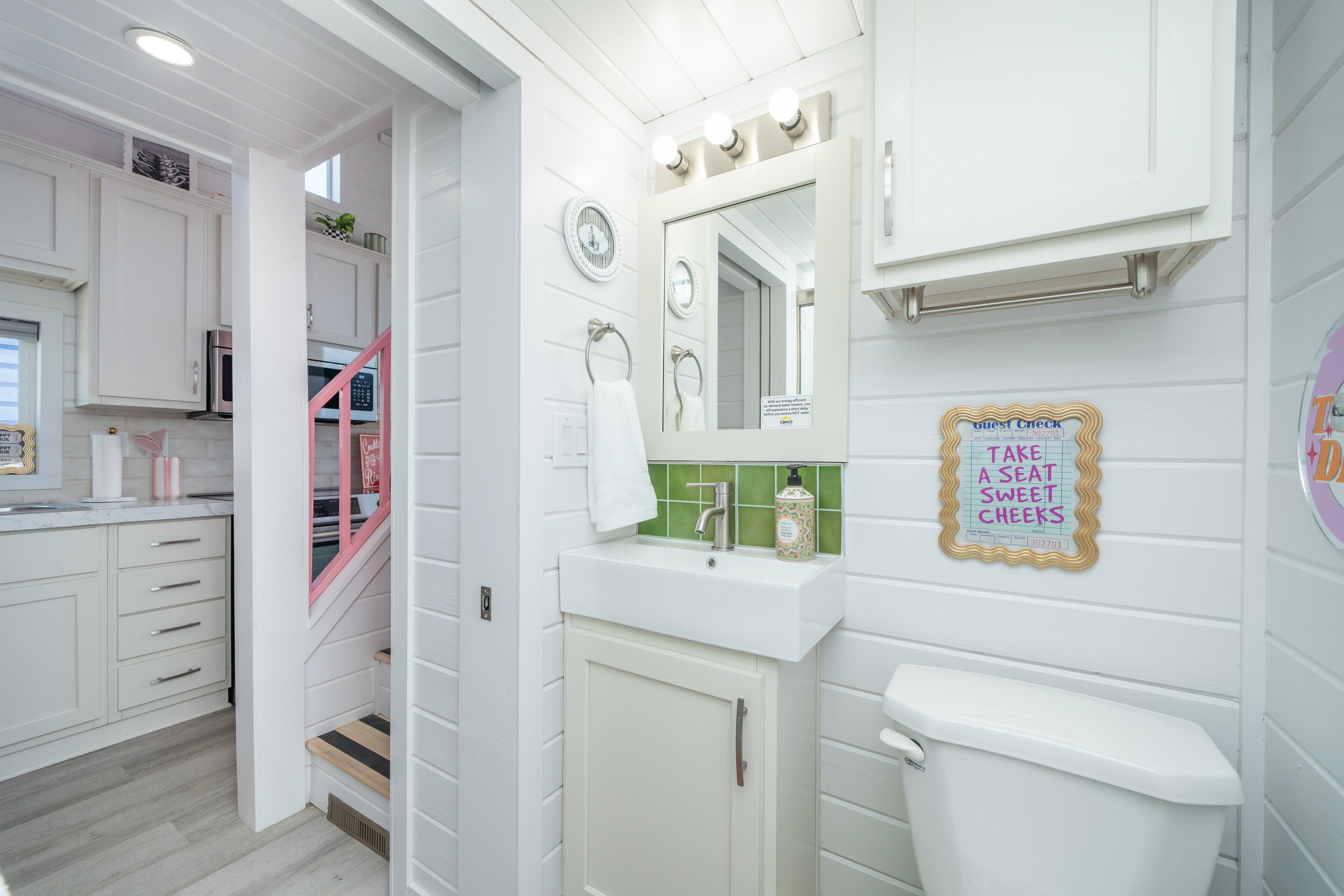 A bathroom with a white sink, mirror, and toilet. There is a sign on the wall that says "TAKE A SEAT SWEET CHEESE."