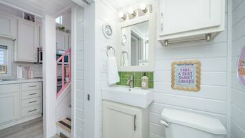 A bathroom with a white sink, mirror, and toilet. There is a sign on the wall that says "TAKE A SEAT SWEET CHEESE."
