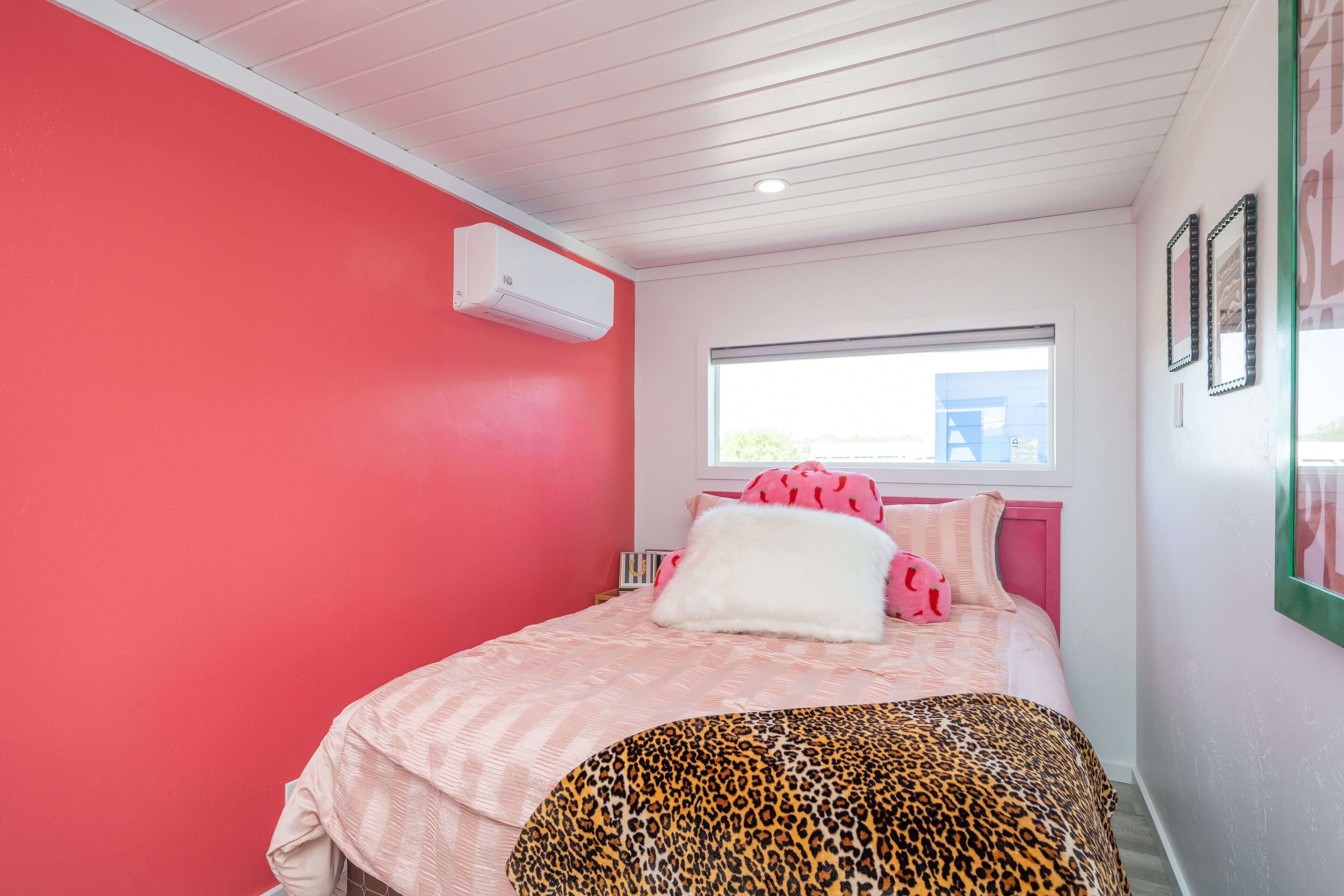 A bedroom with a pink wall, a window, a bed with pink bedding, a leopard print throw, and a white air conditioner.