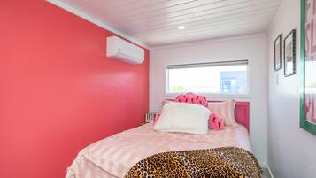 A bedroom with a pink wall, a window, a bed with pink bedding, a leopard print throw, and a white air conditioner.