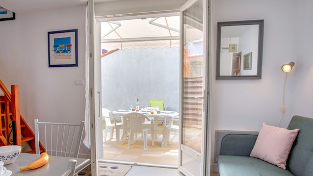 Une pièce avec une porte-fenêtre coulissante donnant sur un espace repas extérieur, un canapé gris avec un coussin rose, une table blanche avec un bol et une banane, et un tableau encadré accroché au mur.