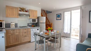Une cuisine compacte avec des meubles en bois, un four à micro-ondes, un réfrigérateur et un lave-linge. On y trouve une table à manger avec des chaises et un petit tapis. Un escalier mène à l’étage supérieur.