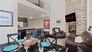 A modern living room with a stone fireplace, leather sofas, a dining area with a wooden table and blue plates, and a kitchen in the background. - 3min Walk to Ski Lifts*Movie Theater*Views (Brian Head)
