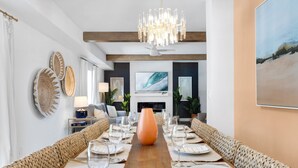Ein langer Esstisch mit einer orangefarbenen Vase in der Mitte, umgeben von gewebten Bänken, in einem modernen Interieur mit einer Chandelieber und Kunstwerken an den Wänden.