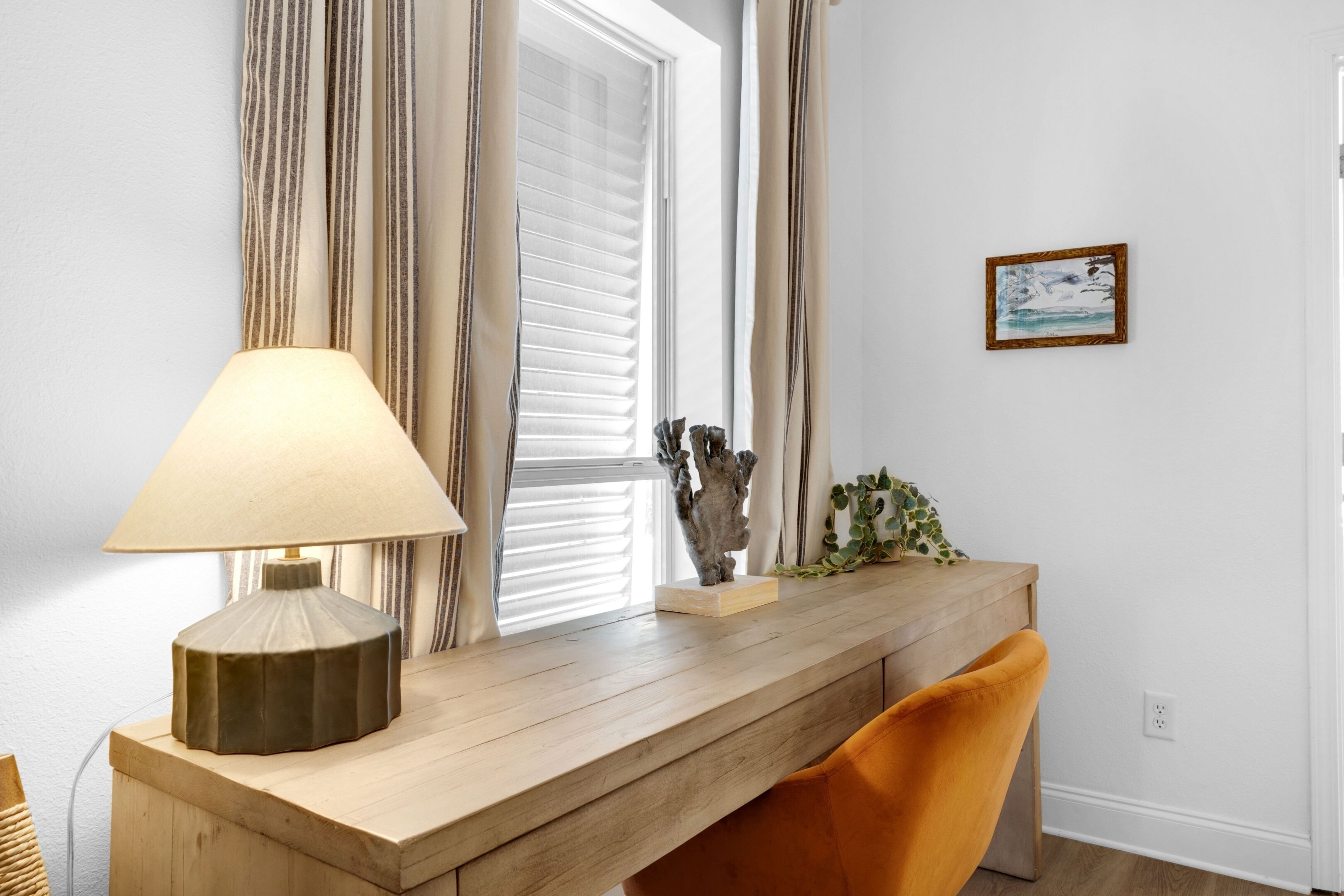 Ein Zimmer mit Schreibtisch aus Holz, einer Lampe, einem Sessel, einem Fenster mit Jalousien und einem gerahmten Bild an der Wand.