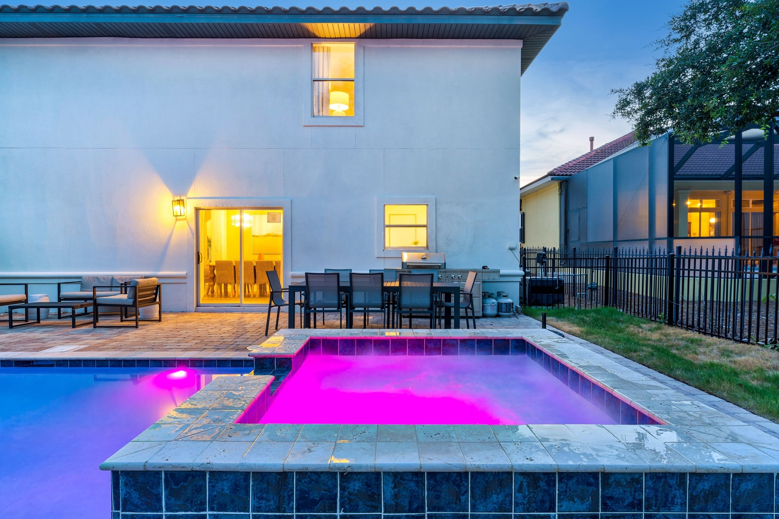 A swimming pool with a tiled edge, a patio area with outdoor furniture, and a building with a lighted entrance.