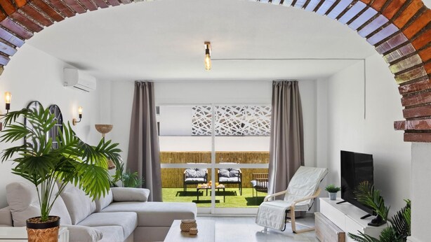 A modern living room with a brick archway, a sofa, a coffee table, a TV, and a view of a garden through large windows. - Erisa 1-29. Terrace, Swimming Pool, Second line of beach (Torremolinos)