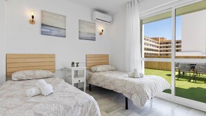 A modern hotel room with two beds, a small bedside table, wall-mounted air conditioning, and a large sliding glass door leading to an outdoor area with a view of a building and a grassy lawn. - Erisa 1-29. Terrace, Swimming Pool, Second line of beach (Torremolinos)