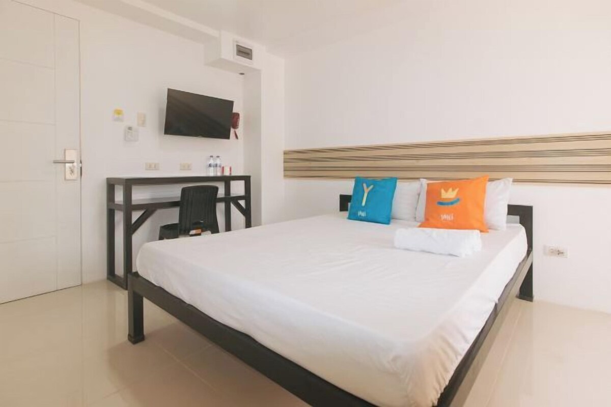 Um quarto com uma cama, uma escrivaninha, uma cadeira, uma TV presa na parede e uma porta.