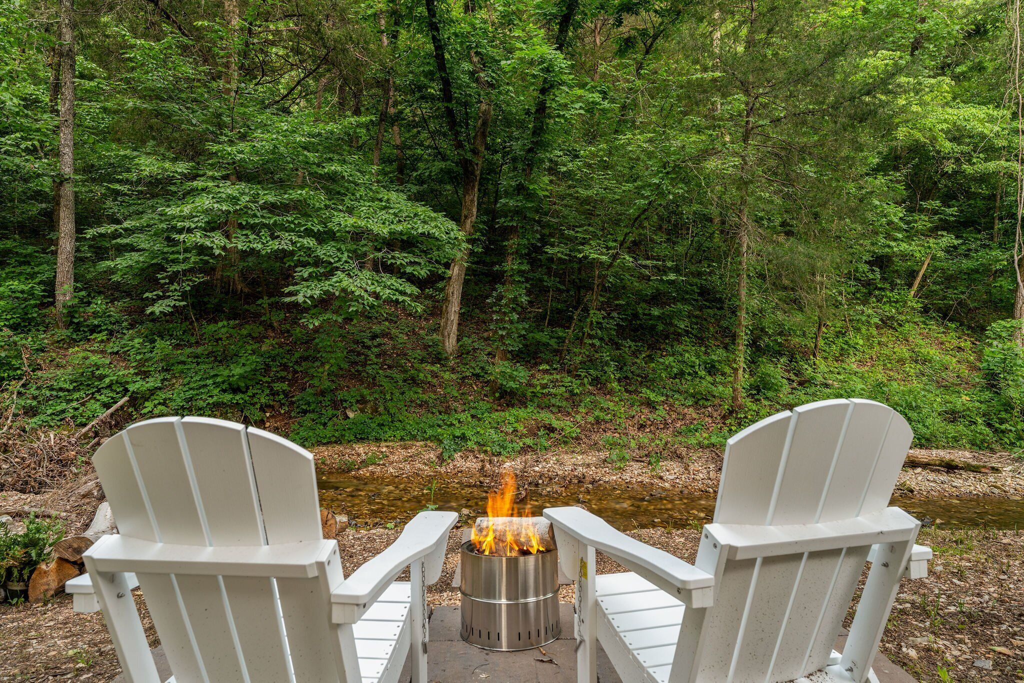 Two white Adirondack chairs facing a campfire in a forested area with a stream in the background.