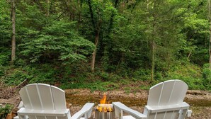 Two white Adirondack chairs facing a campfire in a forested area with a stream in the background. - Stylish Creekside Escape | Pool + Hot Tub (Blue Eye)
