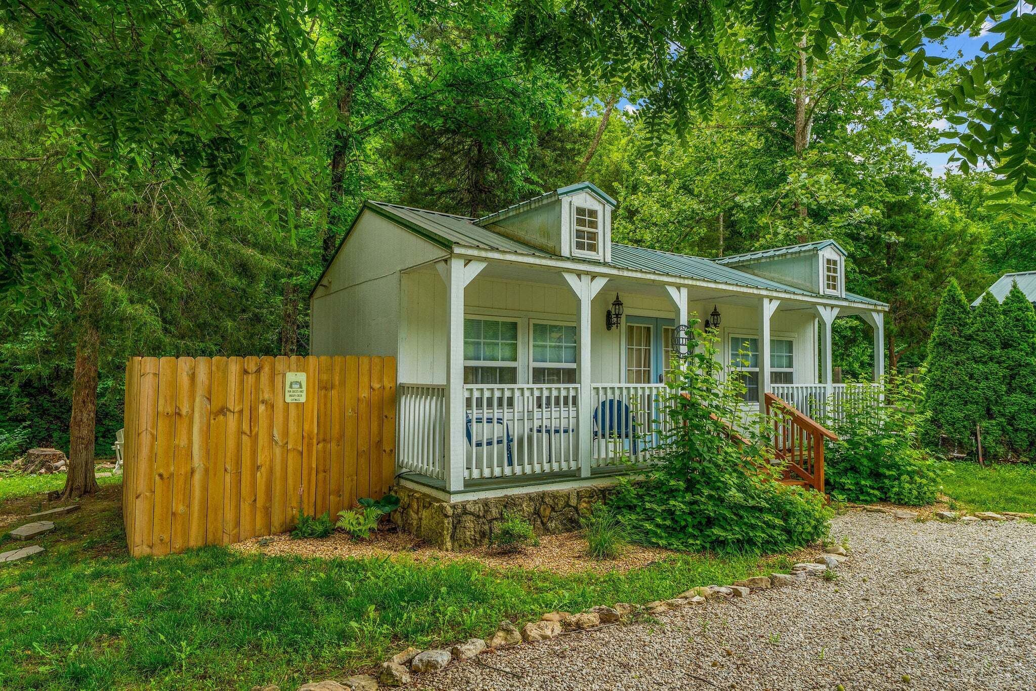 A small, single-story cabin with a porch, surrounded by dense forest.