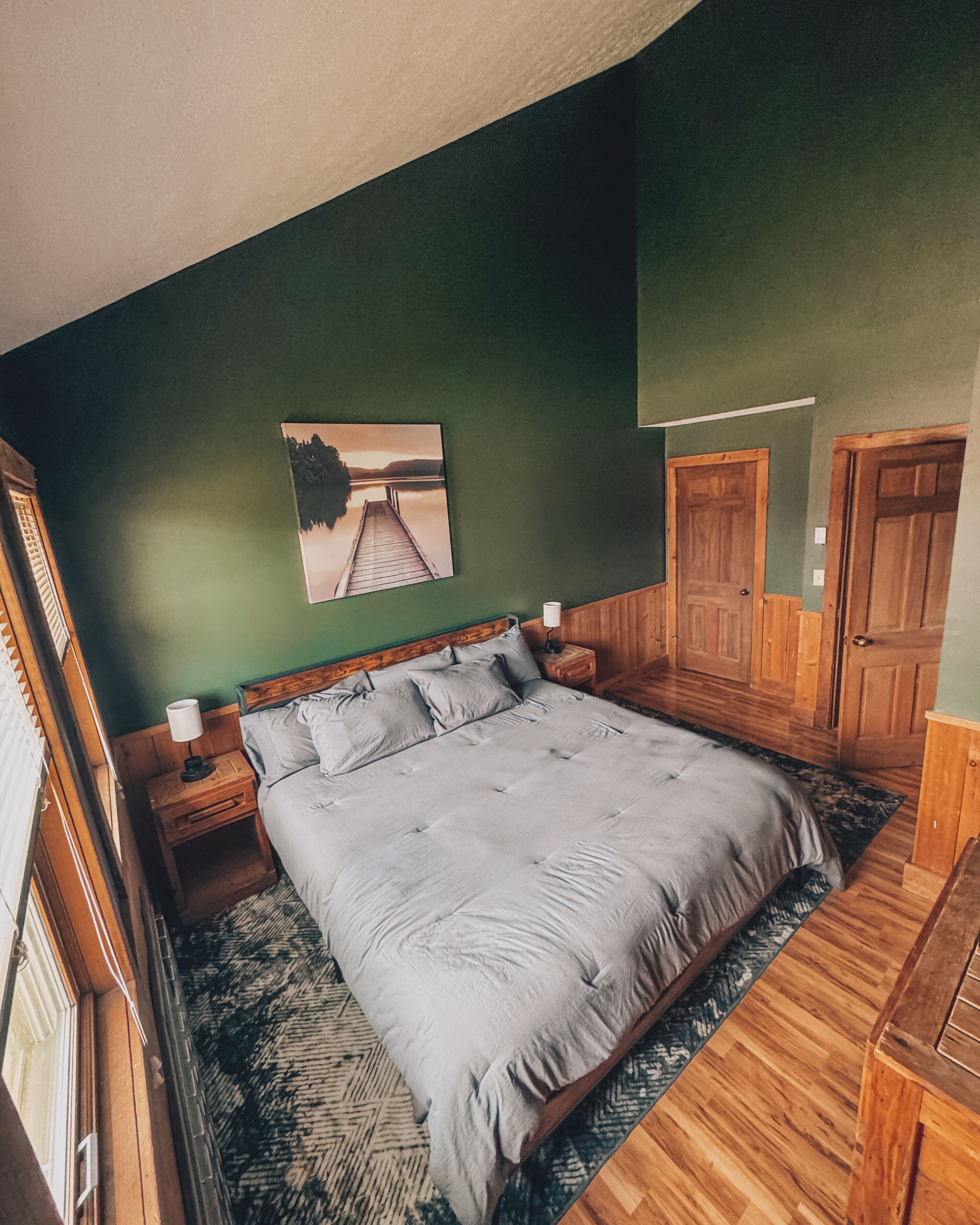 A bedroom with a large bed, wooden headboard, a nightstand with a lamp, a painting on the wall, and a door.