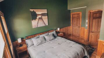 A bedroom with a large bed, wooden headboard, a nightstand with a lamp, a painting on the wall, and a door.