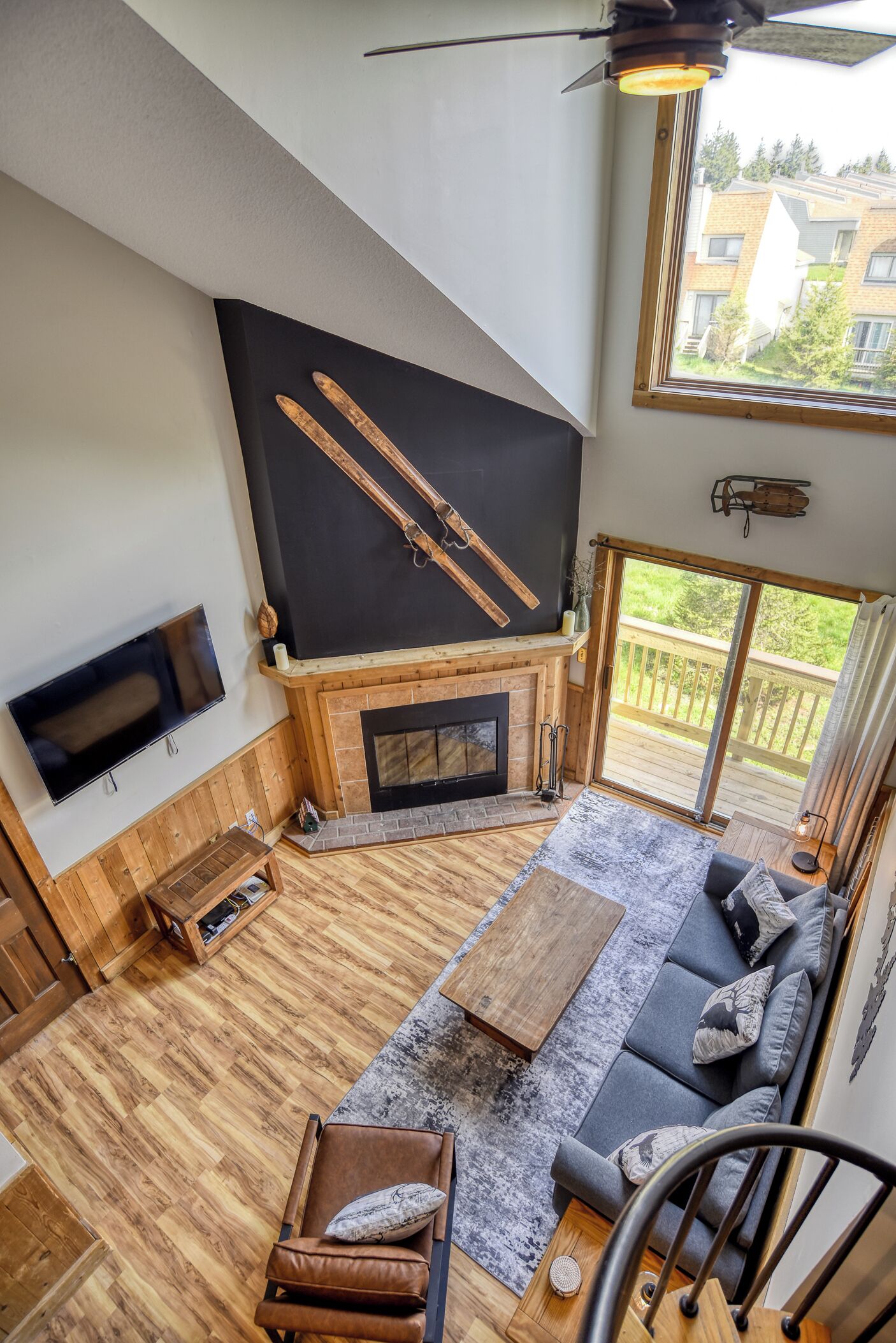 A modern living room with a fireplace, wooden flooring, a large TV, and a balcony with a view of buildings and greenery.