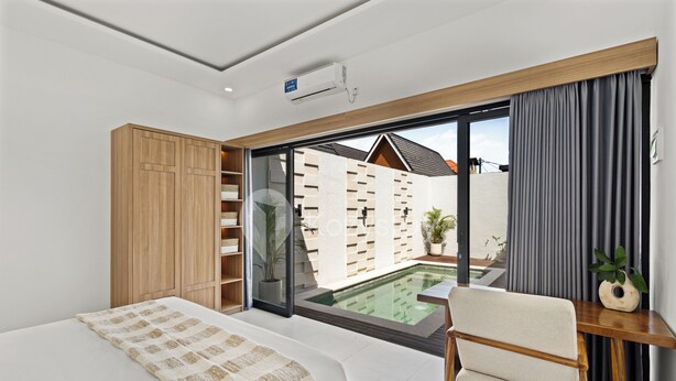 A bedroom with a bed, a wooden wardrobe, a desk with a chair, a window with curtains, and a view of a swimming pool. - Navaya Villa by Kozystay - Seminyak (Seminyak)