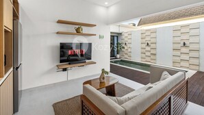 A modern living room with a flat-screen TV, wooden shelves, a sofa, and a pool area visible through the glass wall. - Navaya Villa by Kozystay - Seminyak (Seminyak)
