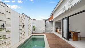 A modern outdoor pool area with a wooden deck, a white wall with a geometric pattern, and a seating area with a table and chairs. - Navaya Villa by Kozystay - Seminyak (Seminyak)