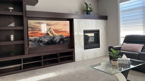 A modern living room with a large flat-screen TV mounted on a dark wooden wall, a fireplace, and a glass coffee table with a tissue box.