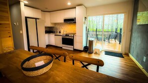 Una cocina moderna con armarios blancos, una estufa y un microondas. Hay una mesa de comedor con una cesta y bancos. Una puerta corrediza de vidrio da a una terraza exterior con sillas.