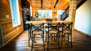 Interior acogedor de una cabaña de madera, con una mesa central, cuatro sillas, chimenea y una escalera.