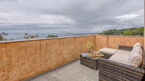 Ein Balkon mit Korbmöbeln, einem kleinen Tisch mit einer Vase und einem Tablett, und Blick aufs Meer.