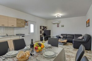 Eine moderne Küche mit weißen Schränken, einem Esstisch gedeckt mit Tellern, Gläsern und einer Obstschale. Im Wohnbereich befindet sich eine schwarze Ledersofa und ein Fernseher.