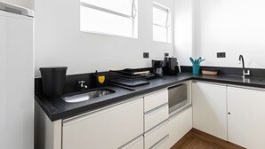 A modern kitchen with white cabinets, a black countertop, a sink, and a coffee maker. - ARK Houses - Studio Completo no Gonzaga (Santos)