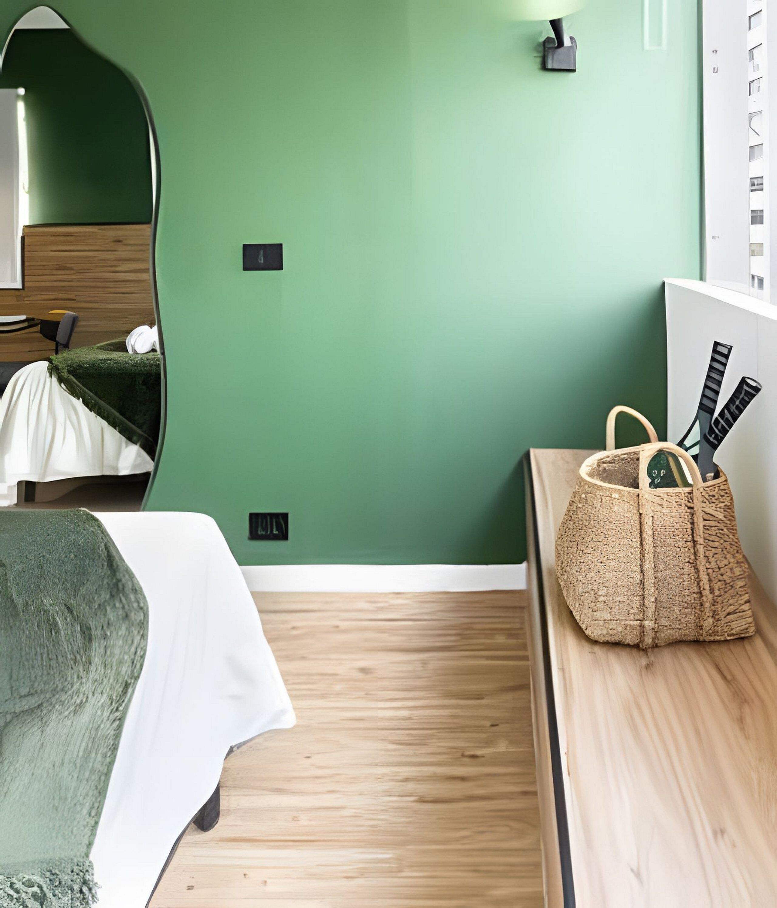 Een kamer met een groene wand, een bed met een witte dekbedovertrek, een houten nachtkastje en een geweven tas met bestek.