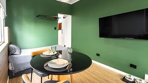 A compact living space with a dining area, a flat-screen TV mounted on the wall, and a small kitchenette. - ARK Houses - Studio Completo no Gonzaga (Santos)