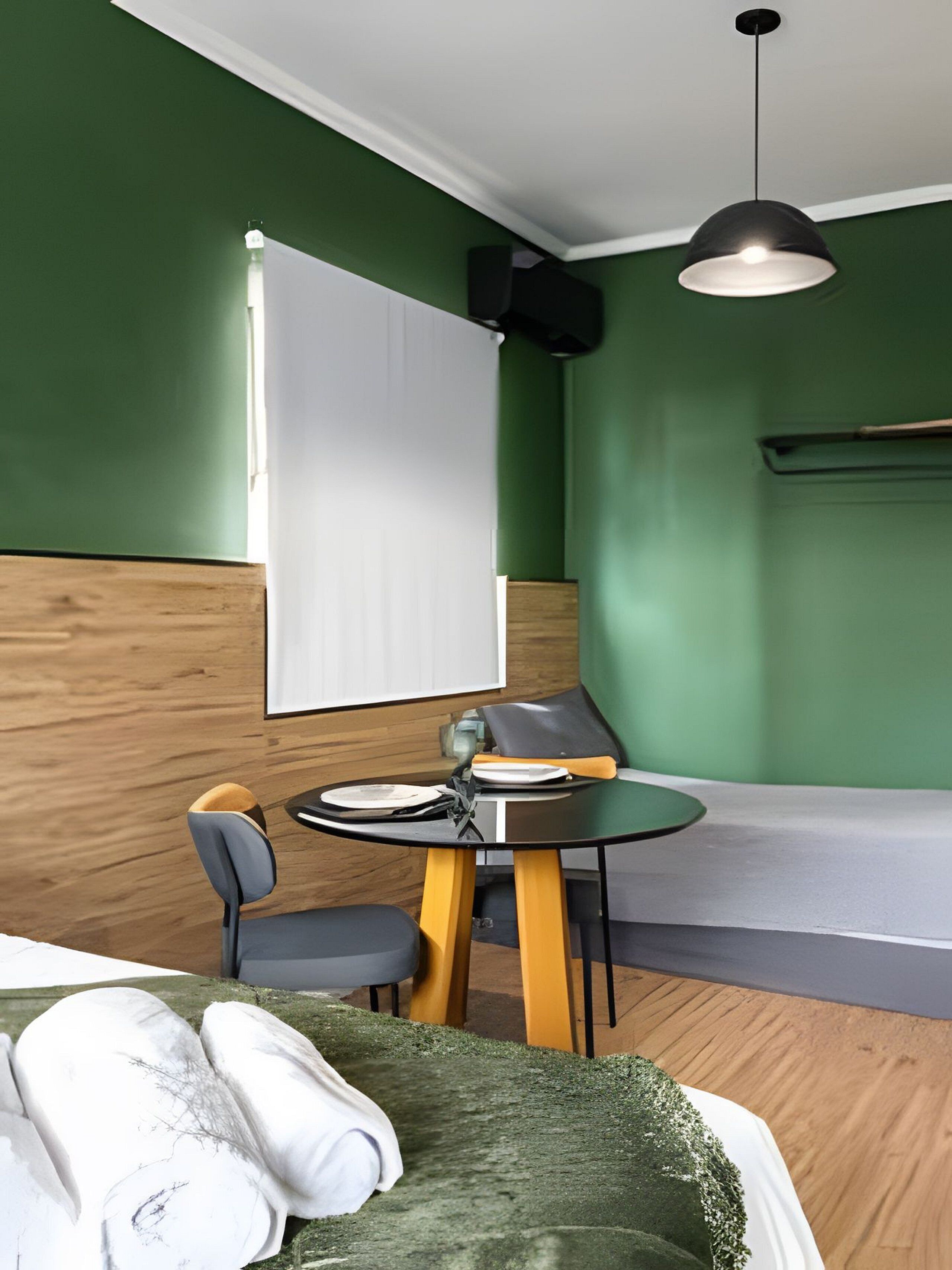 A modern room with green walls, a wooden accent wall, a bed with white linens, a round table with chairs, and a hanging light fixture.