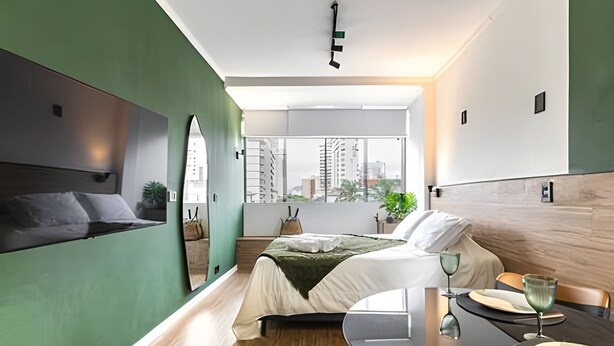 A modern hotel room with a bed, a dining table, and a view of the city. - ARK Houses - Studio Completo no Gonzaga (Santos)